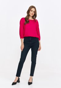 Fuksjowa bluzka z rękawami o długości trzech kwart, połączona z dopasowanymi ciemnymi dżinsami i czarnymi czółenkami z wąskim noskiem. Jednolity tło.