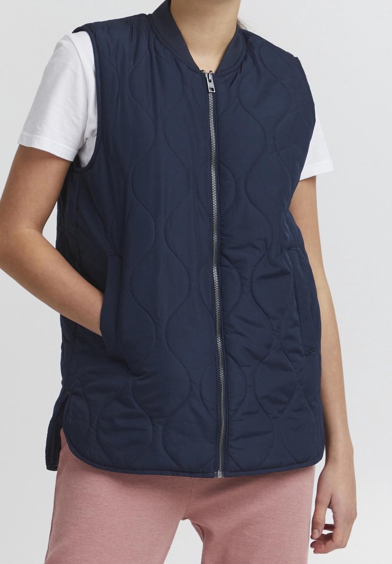 Veste matelassée bleu marine avec fermeture éclair à l'avant, poches latérales et col côtelé. Le matériau semble lisse et léger, conçu pour être porté en superposition.