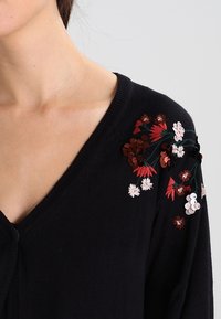 Schwarze Strickjacke mit floralem Stickereimotiv und Pailletten in Rot, Pink und Weiß. V-Ausschnitt-Design mit weichem Stricktextil.