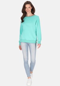 Felpa turchese con colletto tondo e polsini a coste, abbinata a jeans skinny azzurri e sneakers bianche. Il modello si posa in posizione eretta.