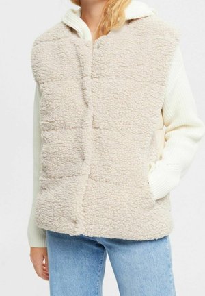 Kvinde iført cremefarvet, tekstureret fleecevest over hvid strikket sweater og lyseblå jeans, hænder i vestens lommer.
