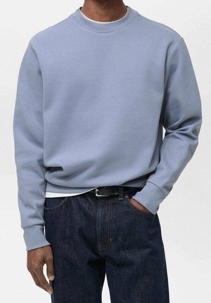 Mann trägt hellblauen Pullover mit Rundhalsausschnitt, dunkelblaue Jeans mit schwarzem Gürtel, eine Hand in der Tasche, vor hellem, einfarbigem Hintergrund.