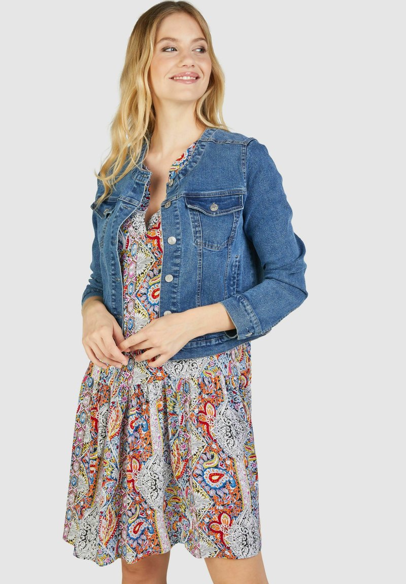 Denim jakna srednje plave boje, s klasičnim gumbima i dva prednja džepa, u kombinaciji s šarenom, uzorkastom haljinom.