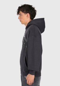 Ung mand med krøllet hår iført en mørk, oversized hættetrøje med et grafisk print, stående i profil mod en ensfarvet baggrund.