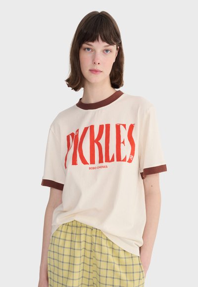 Jeune femme aux cheveux bruns courts portant un t-shirt crème avec des bordures brunes et le texte rouge "PICKLES", associé à un pantalon à carreaux jaunes.