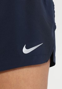 Gros plan sur une personne portant un short de sport bleu foncé avec un logo Nike Swoosh blanc réfléchissant près de l'ourlet.