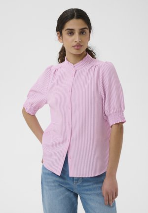 Frau mit dunklem Haar, die eine pink gestreifte Bluse mit Knöpfen und aufgepufften kurzen Ärmeln sowie blaue Jeans trägt, steht vor einem weißen Hintergrund.