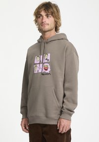 Sudadera marrón con bolsillo delantero, que presenta un diseño gráfico con cuatro rostros de dibujos animados sobre un fondo rosa, marcada con "Volcom" en la parte inferior.