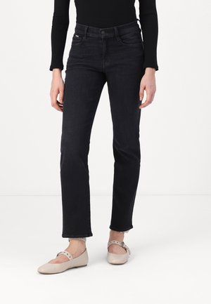 Kobieta ubrana w czarne jeansy o prostych nogawkach, czarną bluzkę z długim rękawem oraz beżowe baleriny typu Mary Jane z paskami wokół kostek, stojąca na białej podłodze.