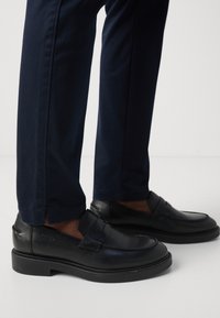 Mocassins en cuir noir avec un bout arrondi et des détails de sangle. Texture lisse, accents cousus et semelle en caoutchouc plate et basse. Le pantalon est bleu marine.