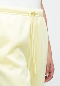 Gaiši dzelteni treniņbikses, kas izgatavotas no mīksta auduma, ar elastīgu jostasvietu un auklu. Tām ir brīvs piegriezums un teksturēta virsma.