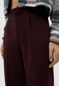 Gros plan sur une personne portant un pantalon taille haute de couleur bordeaux foncé et une veste à carreaux laissant apparaître son ventre et sa main droite.