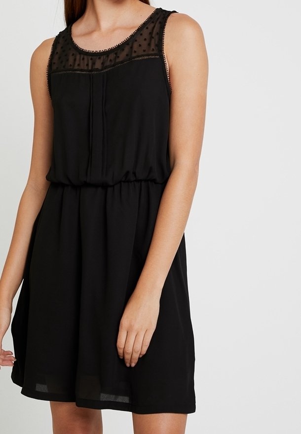 Robe noire sans manches avec empiècement en tulle à pois, encolure ronde, taille cintrée et détail plissé à l'avant portée par une femme debout.