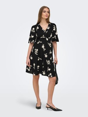 Vrouw draagt een zwarte jurk tot op de knie met witte bloemmotieven, korte mouwen en zwarte slingbackhakken, ze houdt een kleine zwarte tas vast.