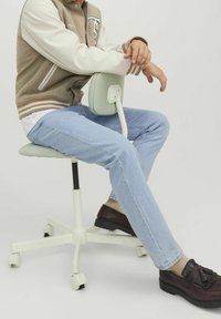 Kontorsstol med en mjuk, mintgrön stoppad sits och ryggstöd, vit bas och hjul. Person klädd i en beige och vit jacka, ljusa jeans, bruna skor.