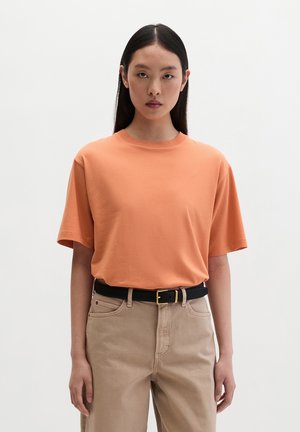 Jonge vrouw met lang donker haar, een losse perzikkleurige T-shirt die in beige broek met een zwarte riem is gestopt, staand tegen een effen achtergrond.