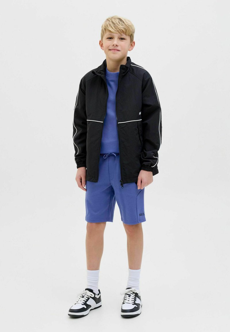 Giacca nera con cerniera e dettagli bianchi, felpa blu e pantaloni corti blu. Sneakers in bianco e nero, calzini bianchi alla caviglia. Stile semplice e informale.