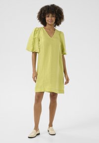 Lime zaļš īss kleita ar V veida kakla izgriezumu, pūkainām piedurknēm un teksturētu materiālu. Saskaņota ar gaišiem mokasīniem.