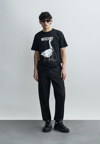 Juoda Moschino marškinėliai su ančiuko grafika ir taškuotu varlyčiu. Derinama su juodomis kelnėmis ir juodais sportbačiais. Modelis dėvi akinius nuo saulės.