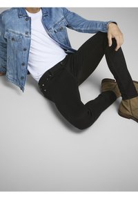 Chaqueta de mezclilla en azul claro, camiseta blanca, jeans skinny negros y botas de ante marrón claro hasta el tobillo. Modelo sentado, con la mano apoyada en la rodilla.