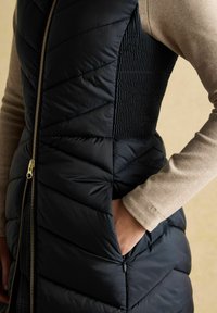 Tuvplānā attēlā ir redzama persona, kas valkā melnu džaku ar quilted struktūru un zelta rāvējslēdzēju, kā arī bēša garām piedurknēm topu, ar roku ievietotu džaka kabatā.