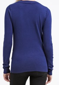 Vrouw in een aansluitende donkerblauwe trui met lange mouwen en zwarte broek, weergegeven van achteren tegen een witte achtergrond.