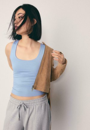 Donna con capelli neri corti che indossa una canottiera blu, pantaloni della tuta grigi e si sta togliendo una giacca marrone con la mano destra.