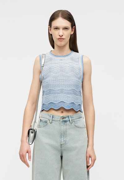 Γυναίκα που φοράει αμάνικο μπλε μπλουζάκι με σχέδιο, ανοιχτόχρωμα τζιν ψηλόμεσα και μια μικρή ασημί τσάντα ώμου.