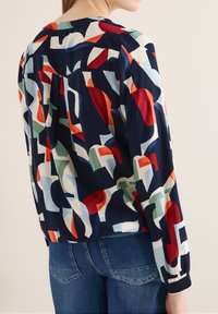 Femme portant un chemisier à manches longues avec un motif abstrait multicolore et un jean bleu foncé, vue de dos.