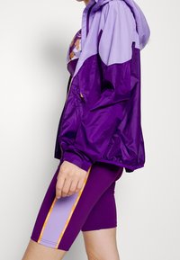 Veste coupe-vent violette avec capuche lilas, dotée de manchettes élastiques et de poches zippées. Assortie à un short de vélo violet avec des accents sur les panneaux latéraux.
