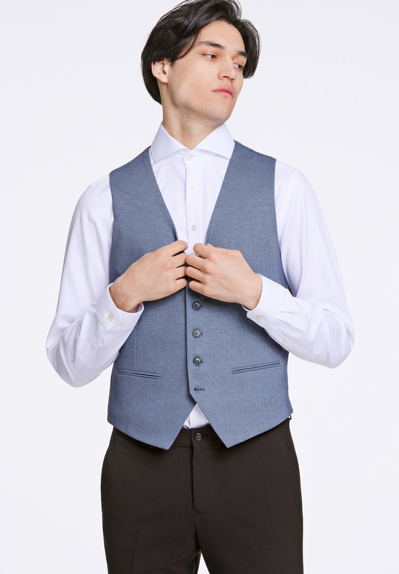 Blå rutigt väst av texturerat tyg. Har fem knappar, två framfickor och en notchkrage, bärs över en vit skjorta.