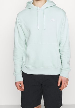 Mann trägt hellblauen Nike-Hoodie mit Fronttasche und schwarzen Shorts, steht vor einfachem weißen Hintergrund.