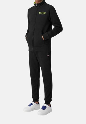 Personne portant un survêtement noir Champion avec une veste zippée et un pantalon assorti, associé à des baskets blanches avec des lacets bleus.
