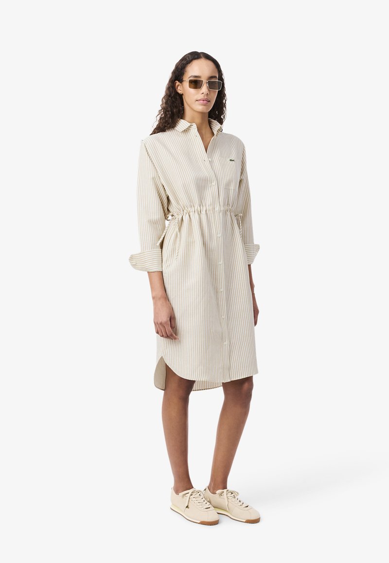 Frau trägt ein beige-weiß gestreiftes Hemdkleid mit geraffter Taille, beige Turnschuhe und rechteckige Sonnenbrille, steht vor weißem Hintergrund.