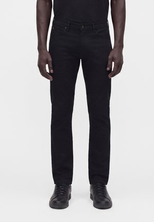 Hombre que lleva pantalones negros ajustados y zapatos negros con cordones, de pie frente a un fondo blanco.