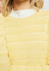 Vaaleankeltainen neulepaita, jossa on vaakasuorat raidat ja avoin kirjoneulekuviointi. Pehmeä koostumus, pyöreä pääntie ja pitkät hihat.