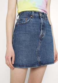 Falda de mezclilla, azul oscuro, ligeramente descolorida. Presenta una cintura alta, cinco bolsillos, botón metálico y dobladillo sin terminar. Textura suave.