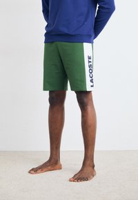 Grønne shorts med hvit sidestropp som viser "LACOSTE" i marineblå. Mykt, teksturert stoff. Modell har på seg en marineblå hettegenser, barføtt.
