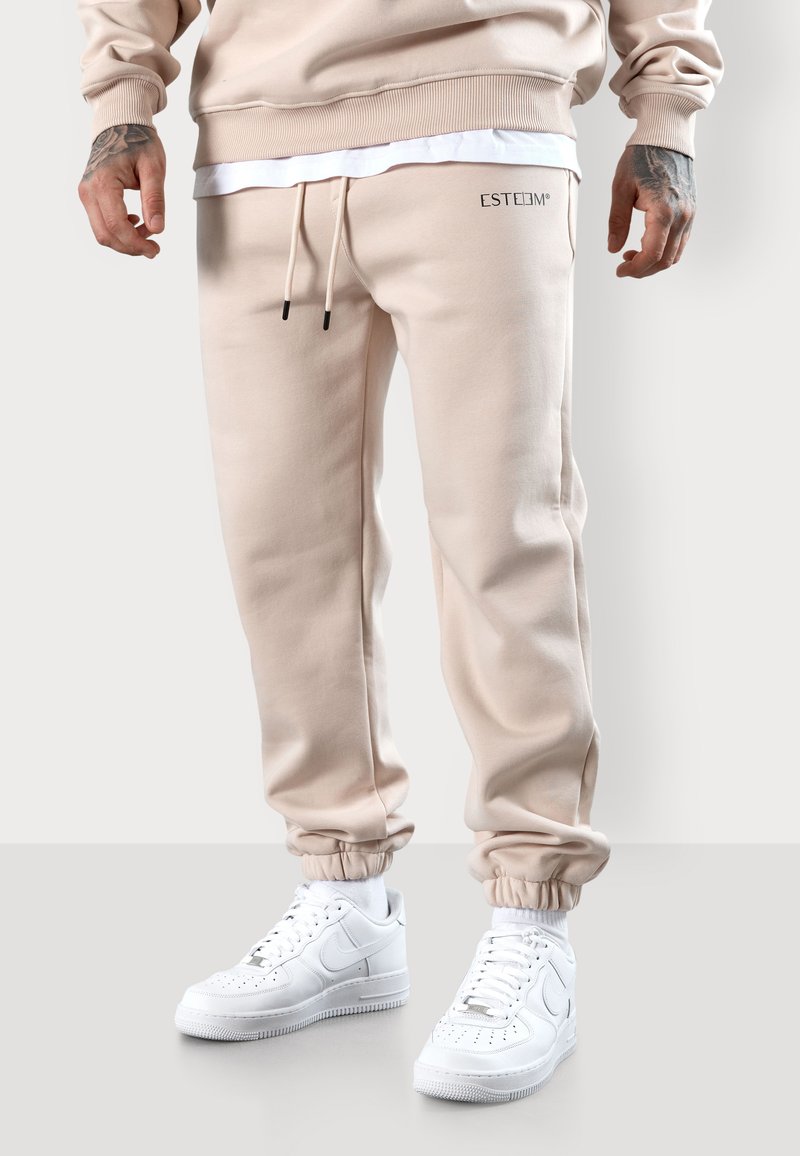 Beige Jogginghose aus weichem Stoff mit elastischen Bündchen, verstellbaren Kordelzügen und einem Logo-Detail, kombiniert mit weißen Sneakern.