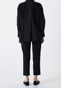 Persona che indossa una maglietta nera a maniche lunghe con spalle testurizzate, pantaloni neri su misura e ballerine nere con fibbia dorata, in piedi su uno sfondo bianco.