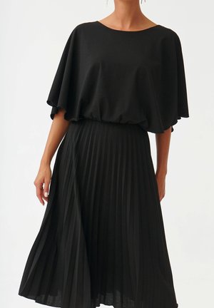 Femme portant un haut à manches cape noir associé à une jupe midi plissée taille haute, sur un fond clair uni.