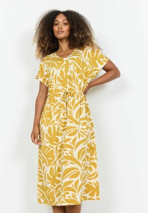 Ženska s kodrastimi lasmi, oblečena v rumeno-belo midi obleko z motivom listov in s trakcem za zavezovanje v pasu, pozira z eno roko na bokih.