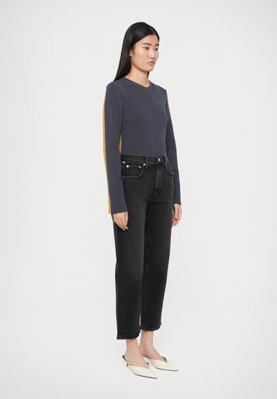 Πλεκτό πουλόβερ σε σκούρο γκρι με ριμπ υφή και μουσταρδί πλευρικές ρίγες, σε συνδυασμό με ψηλόμεσα μαύρα τζιν και λευκές μυτερές μπαλαρίνες.