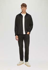 Zwart gestreept shirt met een button-down kraag, gecombineerd met donkere jeans en lichte sneakers. Onder het shirt is een effen wit T-shirt zichtbaar.
