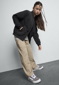 Veste bomber noire avec des poignets côtelés, pantalon cargo beige avec poches latérales, et baskets grises avec des lacets blancs. Le modèle est dans une pose dynamique.