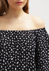 Schulterfreies schwarzes Oberteil mit weißen Blumenmustern, aus strukturiertem Stoff gefertigt und mit einem gerafften Ausschnitt. Kurze Puffärmel sorgen für Volumen.