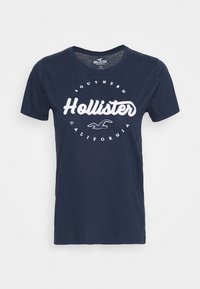 Camiseta de algodón azul marino con cuello en forma de V, con el logo blanco de "Hollister" y un diseño de texto circular que dice "California del Sur". Mangas cortas.