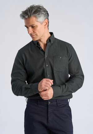 Mann mit grauen Haaren, der ein dunkles Hemd mit Knöpfen und dunkle Hose trägt, schaut nach unten, während er seine linke Hemdmanschette justiert.