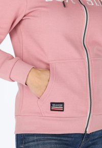 Felpa rosa con zip realizzata in tessuto morbido, dotata di tasche frontali, polsini a coste e una patch con il logo "SEASIDE". Zip con colore a contrasto.