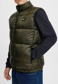 Olivgrüne Steppweste mit hohem Kragen, Reißverschluss und Seitentaschen. Strukturierter, glänzender Stoff mit gesteppten Abschnitten und einem Logo-Patch.